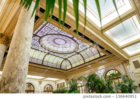 Magnificent stained glass dome ceiling with detailed patterns crowns a historic luxury hotel lobby. Marble columns, arches, and indoor plants complete the refined architectural composition. 136640951