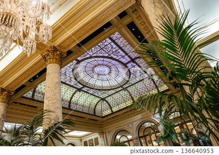 Beautiful stained glass dome ceiling above an opulent hotel lobby with marble columns and lush indoor plants. Classic architecture and intricate details create an atmosphere of elegance and grandeur. 136640953