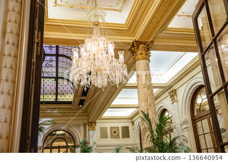 Elegant crystal chandelier hanging from a richly decorated ceiling in a historic luxury hotel. Marble columns, stained glass, and classic architecture create a lavish interior atmosphere. 136640954