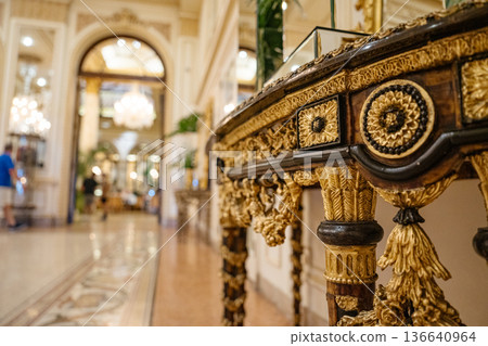 A richly decorated antique table with gold accents and carved details stands in an opulent hotel hallway. The blurred background reveals chandeliers, marble flooring, and classical architectural 136640964