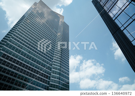Tall glass skyscrapers rise against a bright blue sky in Manhattan, New York City. Reflections of clouds dance on the smooth glass surfaces, emphasizing modern urban architecture. Tall glass skyscrapers rise against a bright blue sky in Manhattan, New York City. Reflections of clouds dance on the smooth glass surfaces, emphasizing modern urban architecture. 136640972