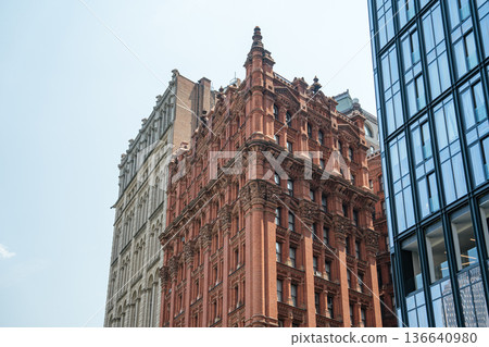 An ornate red brick building stands between historic and modern high-rises in Lower Manhattan. The architectural detail highlights New York City's rich urban heritage. 136640980