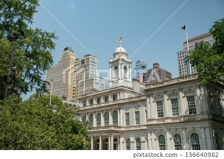 Detailed view of the historic New York City Hall with its iconic white clock tower. Surrounding trees and modern skyscrapers highlight the contrast of old and new architecture. Detailed view of the historic New York City Hall with its iconic white clock tower. Surrounding trees and modern skyscrapers highlight the contrast of old and new architecture. 136640982
