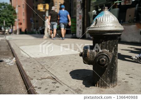 A weathered black fire hydrant stands on a sunlit New York City sidewalk. People walk in the background, giving the street a lively and urban atmosphere. 136640988