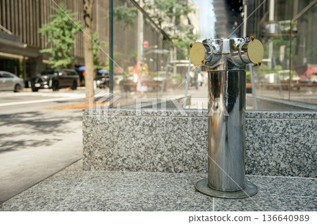 A polished dual fire department connection stands in front of a granite base beside a mirrored glass building. Trees, cars, and blurred street elements form the urban backdrop on a sunny day. A polished dual fire department connection stands in front of a granite base beside a mirrored glass building. Trees, cars, and blurred street elements form the urban backdrop on a sunny day. 136640989