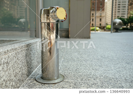 A shiny stainless steel fire department connection stands mounted on a granite sidewalk near a glass building. Urban architecture and blurred buildings appear in the background on a sunny day. A shiny stainless steel fire department connection stands mounted on a granite sidewalk near a glass building. Urban architecture and blurred buildings appear in the background on a sunny day. 136640993