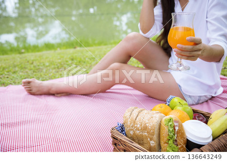 Close up Women picnic in green park hands holding glass of orange juice fresh fruit cool summer. Women drinking sit blanket with Picnic basket bread coffee cup in wood wicker healthy box red Blanket Close up Women picnic in green park hands holding glass of orange juice fresh fruit cool summer. Women drinking sit blanket with Picnic basket bread coffee cup in wood wicker healthy box red Blanket 136643249