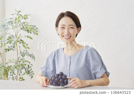 Portrait of a woman eating Kyoho grapes in the living room, looking at the camera Portrait of a woman eating Kyoho grapes in the living room, looking at the camera 136643523