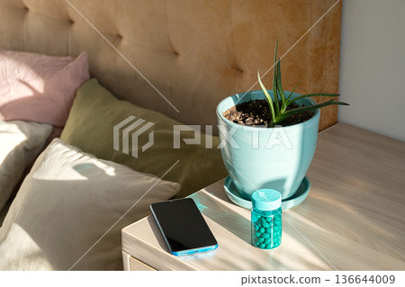 Smartphone, potted plant, and bottle of pills on nightstand beside bed. Smartphone, potted plant, and bottle of pills on nightstand beside bed. 136644009