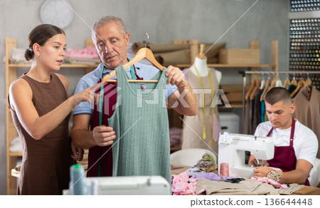 Girl customer examines finished product in presence of mature man tailor, guy sew in background Girl customer examines finished product in presence of mature man tailor, guy sew in background 136644448