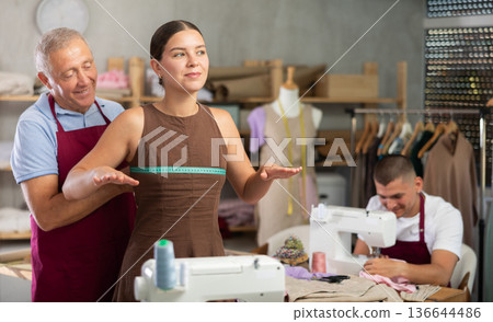 Mature man tailor takes measurements from girl client, guy work with sewing machine Mature man tailor takes measurements from girl client, guy work with sewing machine 136644486