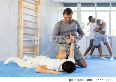 Guy and man are training in gym practicing technique of grabbing arm and lowering opponent to floor Guy and man are training in gym practicing technique of grabbing arm and lowering opponent to floor 136644490