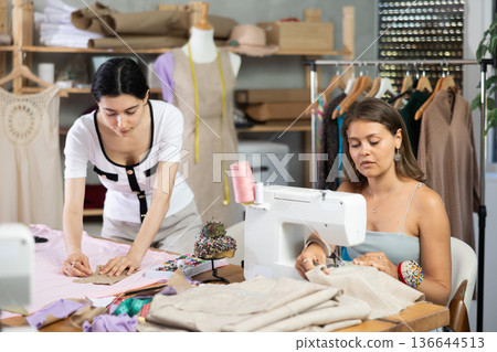 Young seamstress stitching on sewing machine in workshop with colleague Young seamstress stitching on sewing machine in workshop with colleague 136644513