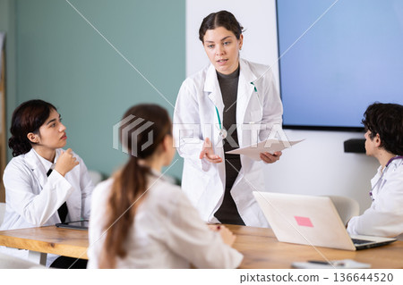 Positive young girl medical student standing with papers in hands near table where sitting colleagues and talking in corporate seminar in office 136644520