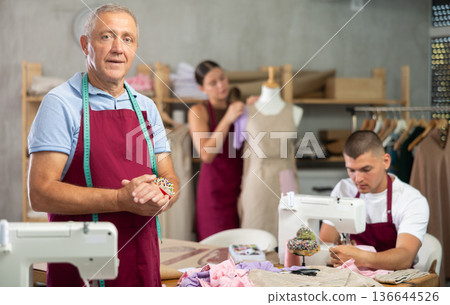 Senior man fashion designer is in office, guy sewing clothes, girl work with mannequin in background Senior man fashion designer is in office, guy sewing clothes, girl work with mannequin in background 136644526