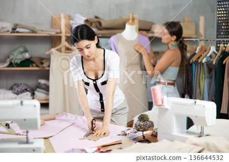 Female tailor cutting fabric while teammate working with mannequin Female tailor cutting fabric while teammate working with mannequin 136644532