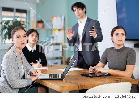 Interested office workers sitting at table discussing ideas and watching presentation during business meeting in office Interested office workers sitting at table discussing ideas and watching presentation during business meeting in office 136644582