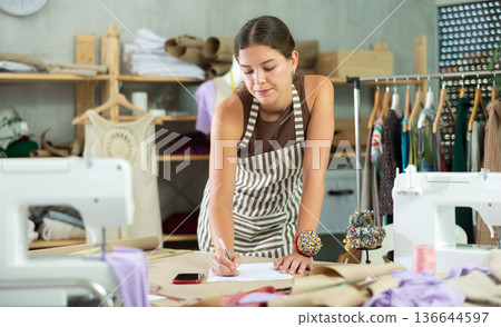 Young seamstress making calculations on paper standing near table Young seamstress making calculations on paper standing near table 136644597