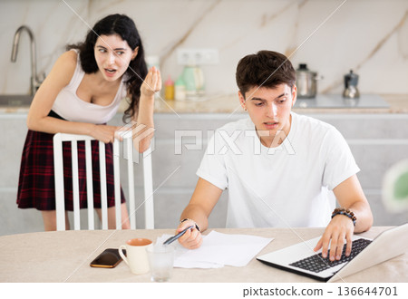 Sad freelancer husband works at home on laptop and listens to her wife reproaches in frustration. Sad freelancer husband works at home on laptop and listens to her wife reproaches in frustration. 136644701