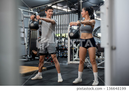Fitness trainer coach man talking teaching asian woman client holding kettlebell exercising in gym. Fitness trainer coach man talking teaching asian woman client holding kettlebell exercising in gym. 136648199