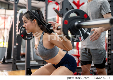 Asian woman client exercising with weight lift barbell while fitness gym trainer coach man teaching. 136648225