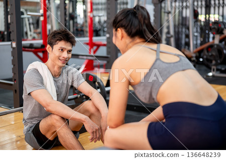 Asian man in sportswear with towel sitting on gym floor taking a rest while talking to woman friend. 136648239