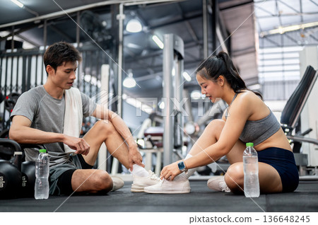 Asian man in sportswear talking to a woman friend tying sneakers's shoelace as sitting on gym floor. Asian man in sportswear talking to a woman friend tying sneakers's shoelace as sitting on gym floor. 136648245