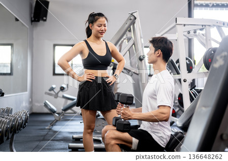 Asian man client holding dumbbell talking to pretty woman fitness trainer after exercising in gym. 136648262