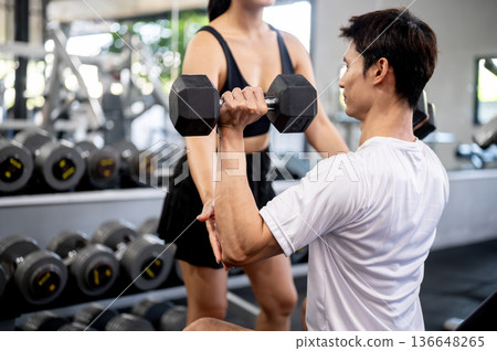 Asian man client holding dumbbell exercising as woman fitness trainer helping fixing posture in gym. Asian man client holding dumbbell exercising as woman fitness trainer helping fixing posture in gym. 136648265
