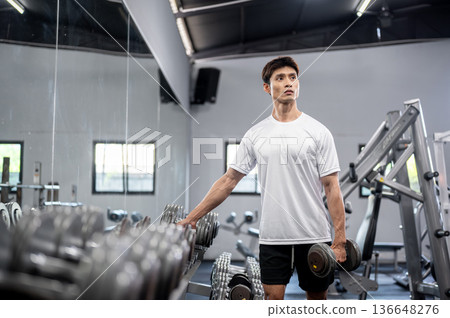 Asian man fitness trainer coach or a client standing and holding a dumbbell while exercising in gym. 136648276