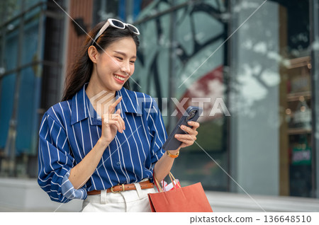 Pretty asian woman holding and looking at phone carrying shopping bags while walking outside a mall. Pretty asian woman holding and looking at phone carrying shopping bags while walking outside a mall. 136648510