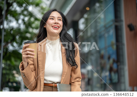 Pretty asian woman office worker holding coffee cup carrying laptop walking outside a shopping mall. Pretty asian woman office worker holding coffee cup carrying laptop walking outside a shopping mall. 136648512