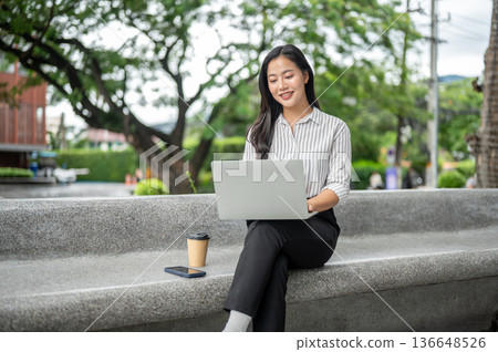 Pretty asian woman office worker looking at laptop sitting aside coffee on stone bench in a garden. Pretty asian woman office worker looking at laptop sitting aside coffee on stone bench in a garden. 136648526