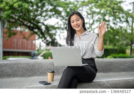 Pretty asian woman office worker waving hand over laptop and coffee sitting on stone bench in garden 136648527