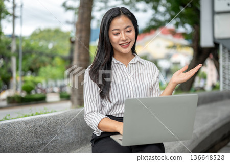 Pretty asian woman office worker explaining with hand over laptop sitting on stone bench in a garden 136648528