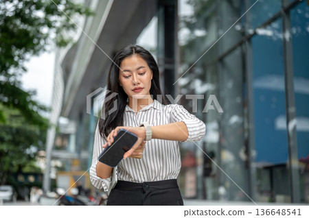 Asian woman office worker holding phone looking at time on watch while walking outside shopping mall 136648541