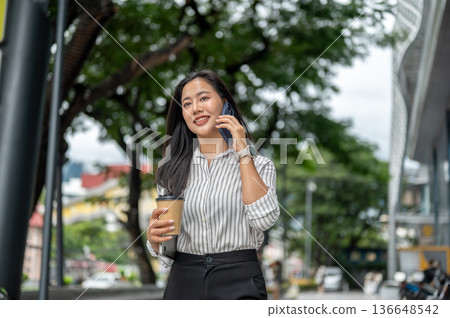 Asian woman office worker holding coffee cup talking on phone while walking outside a shopping mall. 136648542