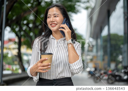 Asian woman office worker holding coffee cup as talking on phone while walking outside shopping mall 136648543