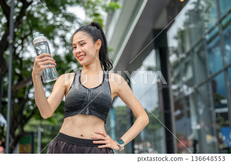Asian woman in sportswear holding looking at water bottle with hand on waist resting after exercise. 136648553