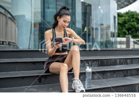 Asian sportswear woman holding phone looking at time watch sitting on stairs resting after exercise. 136648558