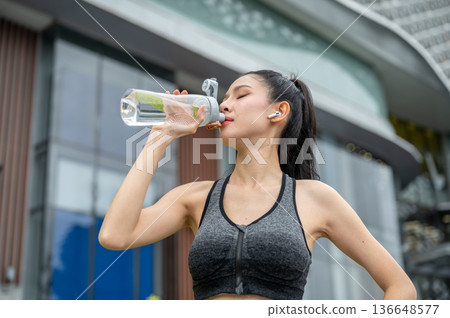 Asian woman in sportswear with earbud closing eyes drinking water after running exercise aside mall. Asian woman in sportswear with earbud closing eyes drinking water after running exercise aside mall. 136648577