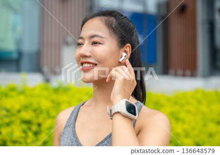 Asian woman in sportswear touching earbud listening to music while running exercising in garden park 136648579