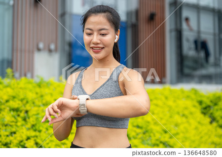 Asian woman in sportswear listening to music looking at smartwatch while running exercising in park. Asian woman in sportswear listening to music looking at smartwatch while running exercising in park. 136648580
