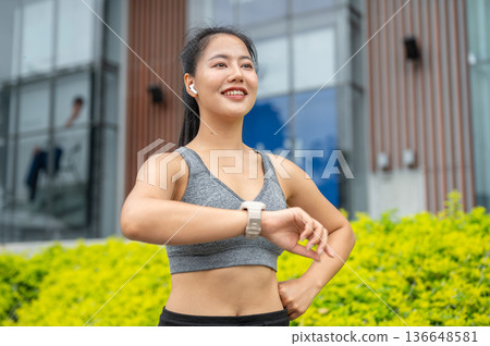 Asian woman in sportswear listening to music looking away from watch while running exercise in park. 136648581