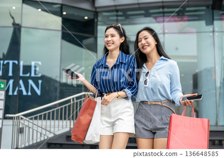 Happy asian woman and friend holding phone carrying shopping bags while walking down the mall's step 136648585