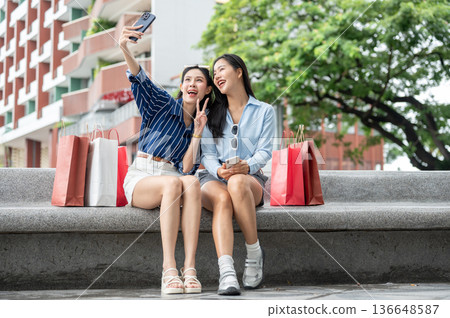 Asian woman friends taking phone picture sitting with shopping bags on bench at mall's garden park. 136648587