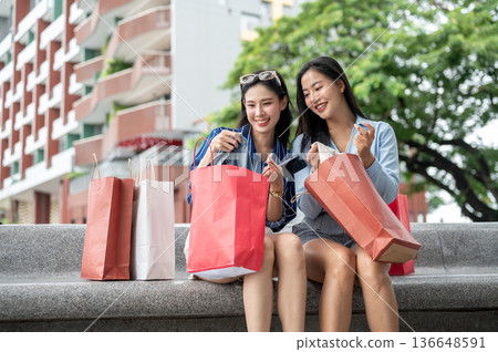 Asian woman showing a cloth in shopping bag to a friend while sitting on bench at mall's garden park Asian woman showing a cloth in shopping bag to a friend while sitting on bench at mall's garden park 136648591