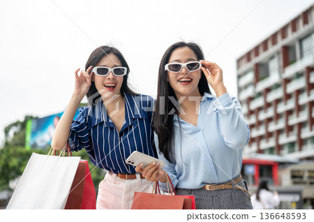 Asian woman friends wearing sunglasses shades carrying shopping bag standing outdoor under clear sky Asian woman friends wearing sunglasses shades carrying shopping bag standing outdoor under clear sky 136648593