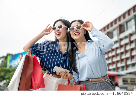 Asian woman friend wearing sunglasses shades carrying shopping bag standing outdoor under bright sky 136648594