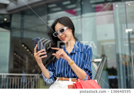 Asian woman with sunglasses looking at phone carrying shopping bag standing in front of a mall plaza Asian woman with sunglasses looking at phone carrying shopping bag standing in front of a mall plaza 136648595